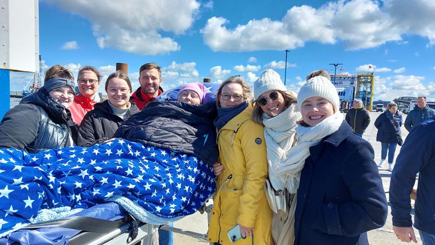 Eine Menschengruppe mit einer Frau auf einer Trageliege in der Mitte, im Hintergrund der Hafen von Dagebüll